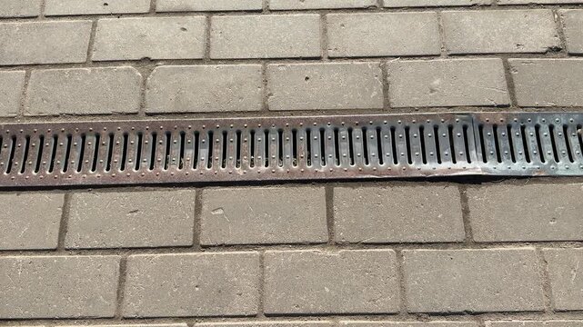 Patterned metal grate on paving stones, close-up view of linear drainage channel embedded in concrete pavers, rusted slats.