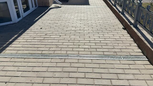 White pipe metal grate on pavement, patterned metal grate covers spillway installed across paving stones on footpath, sunlight.