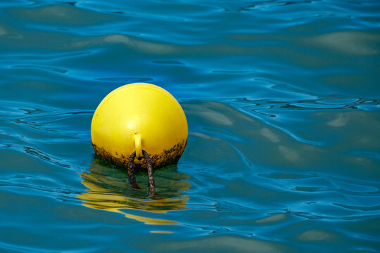 Bright yellow spherical plastic marine buoy floating gently on the surface of deep blue undulating sea water tethered securely to the unseen bottom by an old rope