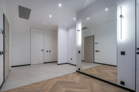 An entryway in a new apartment shows clean walls, a large mirror reflecting light, and a view of closed doors. A wooden floor design adds a unique touch to the space