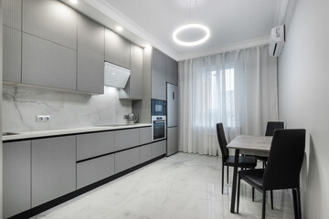 Fototapeta premium A kitchen features grey cabinets and white marble countertops. There is a small dining table with black chairs. Natural light fills the room through the window