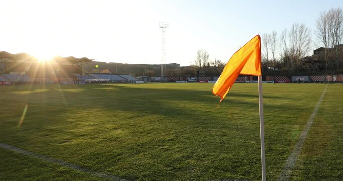 An orange corner flag flutters in the wind on an empty soccer field. The sun shines brightly over the stadium, illuminating the green grass before a football match.