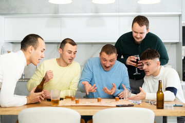 Fototapeta premium Cheerful young adults enjoying beer and wine immersed in board game with twist, where shot glasses of alcoholic drinks replacing traditional game pieces