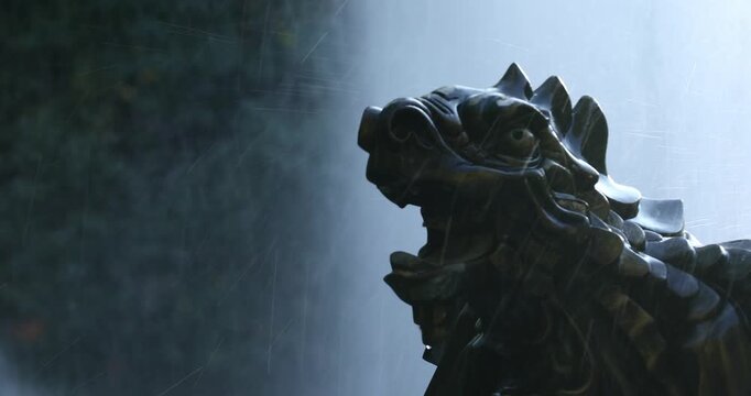 Close-up shot of heavy water pouring over a dark stone statue on a textured wall, with the camera tilting up to reveal a bright, intense spray of water.

