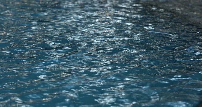Close up of vibrant blue swimming pool water with gentle ripples. Bright sunlight reflects on the moving liquid surface, creating a truly relaxing, calming abstract background texture pattern.

