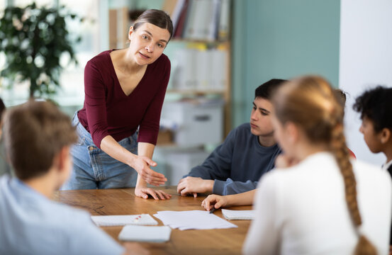 Female teacher gives a task to a group of schoolchildren who are sitting at a table in a classroom