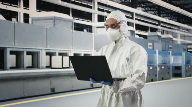 Microchip factory specialist tests silicon wafer batches with laptop software. Technician wearing protective gear monitors semiconductor manufacturing machinery readouts with notebook, camera A