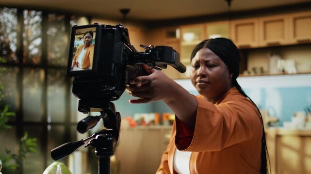 Behind the scenes of African American vlogger adjusting camera on tripod in home kitchen studio. Content creator checks lighting settings and steady framing during preparations, ready for filming.