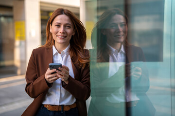 Smiling businesswoman holding smartphone reflecting on glass building