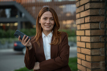 Smiling businesswoman holding phone leaning on brick wall