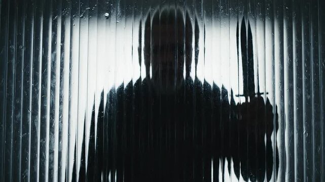 This captures a tense and foreboding scene of a mysterious figure in a black hoodie holding a knife in a dimly lit room with a corrugated metal wall, evoking feelings of fear and anxiety.