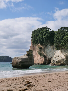 Iconic limestone rock formation at Cathedral Cove, Coromandel Peninsula, New Zealand. Secluded sandy beach with turquoise ocean waves and lush green cliffs under a cloudy blue sky.