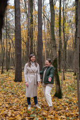 Fototapeta premium Two women talking and laughing during an autumn walk in a forest with yellow leaves and tall trees, natural light and relaxed mood