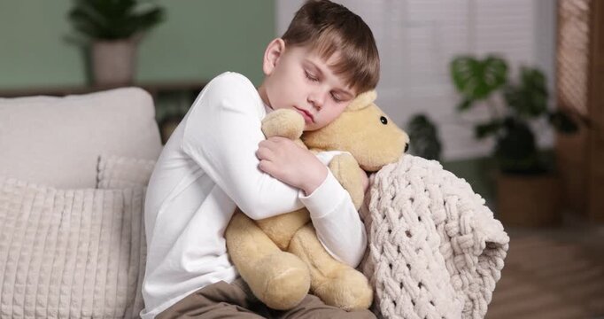 Upset little boy hugging his teddy bear on sofa at home