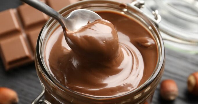 Woman taking tasty chocolate paste with spoon from jar at grey table, closeup