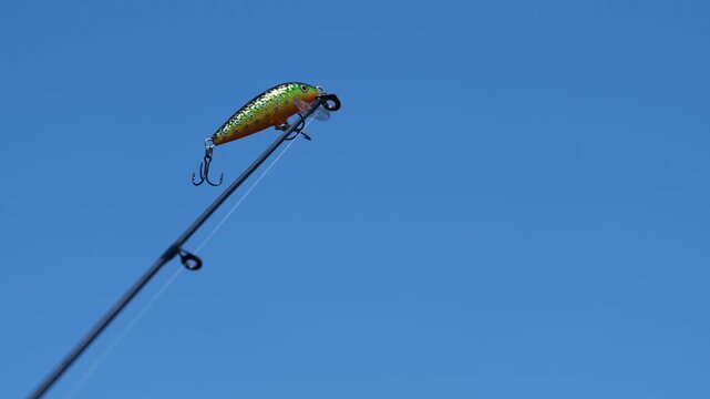 Fishing lure attached to a rod against a clear blue sky, gently vibrating in motion. Artificial lure used in lure fishing, highlighting angling technique, equipment
