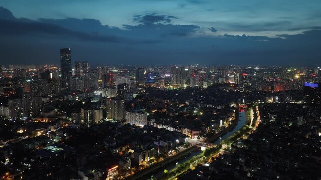 Night aerial view of Hanoi along Lang Street with illuminated traffic trails, glowing skyline, dense neighborhoods, and vibrant urban atmosphere after sunset.