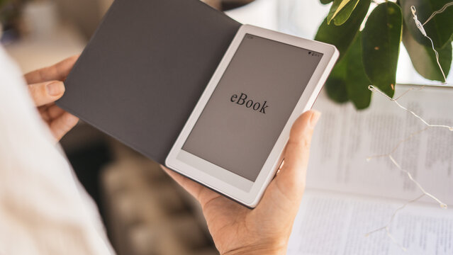 Person reading ebook on digital device near window with houseplants and books. Modern technology meets cozy lifestyle. Concept of home education, relaxation and minimalism