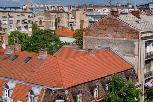 View over dense city rooftops with red tile roofs, chimneys and weathered apartment buildings, mixed old and modern architecture with trees between blocks under bright blue summer sky