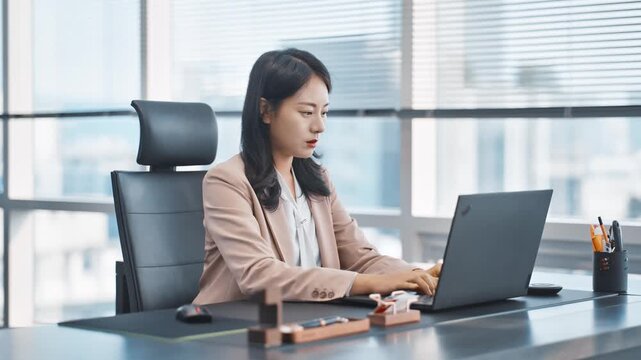 Focused Asian businesswoman diligently working on a laptop in a bright, modern office, embodying productivity and corporate success.