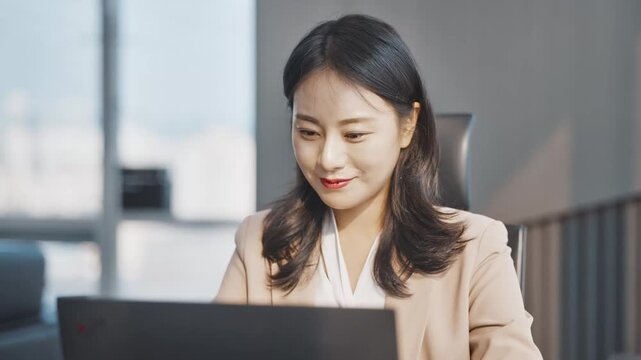 Focused Asian businesswoman working on a laptop in a modern office, diligently engaged in her professional tasks with a dedicated expression.