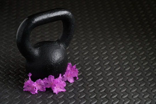 "
Celebrate spring with fitness and healthy lifestyle, black iron kettle bell weight with pink purple azalea flowers on black textured gym floor mat"

