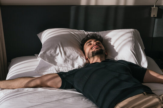 Young man sleeping peacefully in bed, eyes closed, enjoying morning sunlight streaming through the window in a quiet bedroom