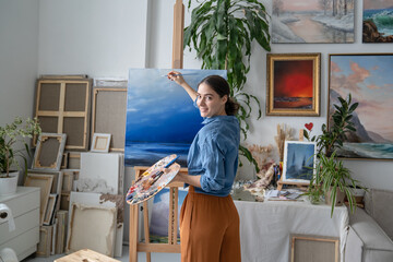 Carefree cheerful woman painter paints picture on easel at studio apartment, art therapy brings positivity and creativeness in everyday life. Smiling female artist enjoying drawing picture on canvas. © DimaBerlin