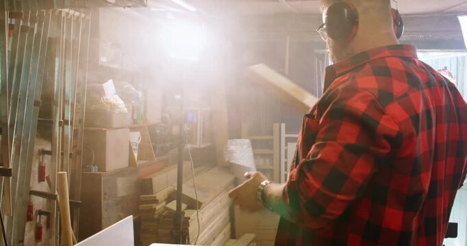Carpenter work. Furniture making. Man putting on protective earmuffs processing wood on thickness planer in bright lamp light interior.