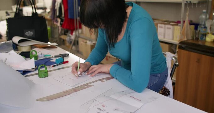 Dressmaking process. Tailor desk. Woman hand writing pencil notes on paper garment patterns creating clothes draft in studio interior.