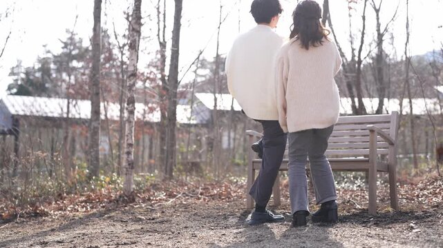 Sunlight glows through bare winter trees. Man holds a child, smiling warmly. Woman stands beside them, content and calm. They pause near a bench, hands joined