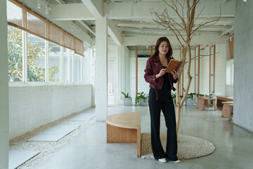 Young woman standing and focusing on reading a book, finding leisure and quiet time in a bright, modern interior space with a dried decorative tree and natural light © MINAE