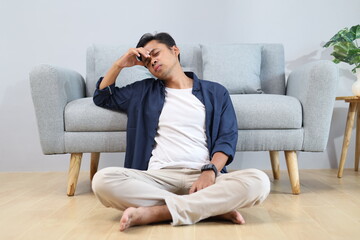 Fototapeta premium A young man sits cross-legged on the floor leaning against the sofa at home, holding his head and closing his eyes, feeling dizzy or having a headache and stress.