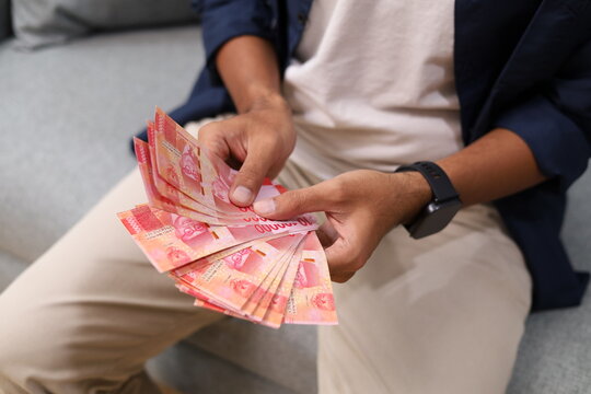 Close up shot of a young man spending the day at home, sitting on the sofa in the living room, counting a wad of rupiah banknotes.