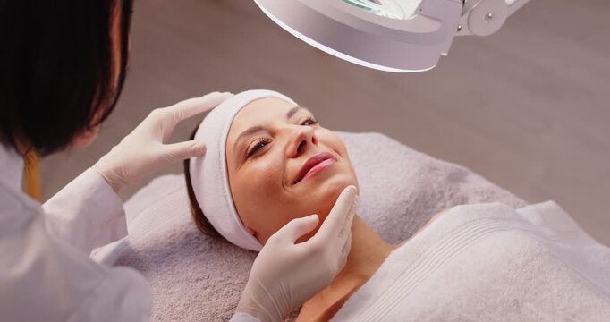 Top view of young woman lying on couch during professional facial skin examination in beauty spa salon. Cosmetologist checking skin condition of female client under lamp before skincare procedure.