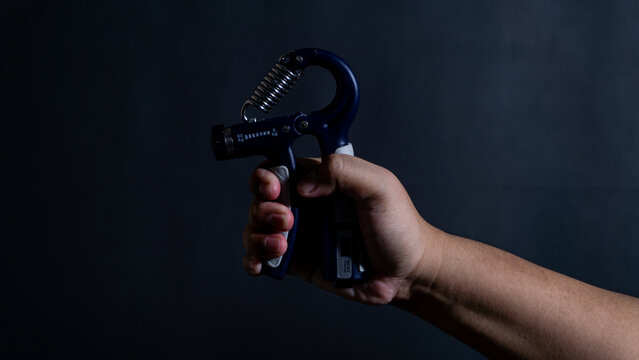 A male hand tightly grips a blue adjustable hand strengthener tool against a solid dark background, showing visible tension in the fingers and arm muscles. Dramatic side lighting highlights