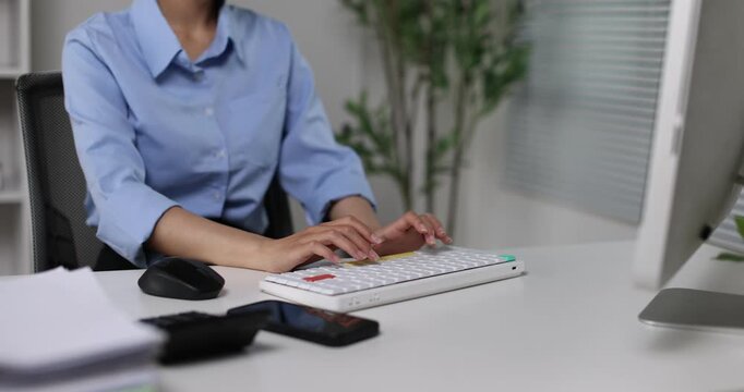 Asian businesswoman working diligently at her desk in a contemporary office setting, performing data entry and managing tasks on a desktop computer, focusing on productivity and professional work