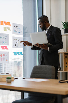 An African American businessman in a suit holds a laptop while pointing at graphs and charts displayed on a glass wall in an office setting