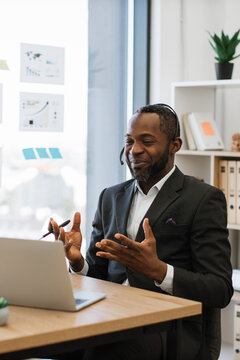 An African American man wearing a suit and headset smiles while gesturing during a video conference call on his laptop