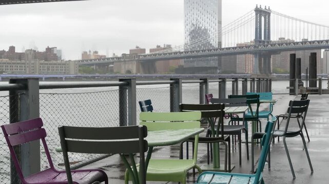 Manhattan Bridge from Brooklyn Bridge park, New York City Dumbo United States. Waterfront promenade, riverside esplanade on rainy foggy day. Empty wet metallic chairs and tables by East river water.