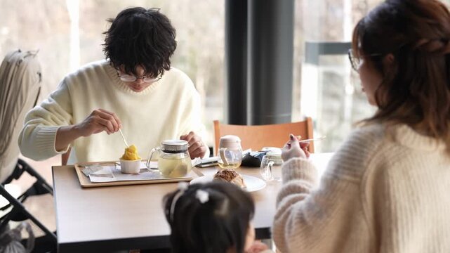 Cozy cafe scene with three people. Soft light fills the room. Person in white sweater eats dessert. Another person watches with quiet interest. Child sits nearby, focused on food