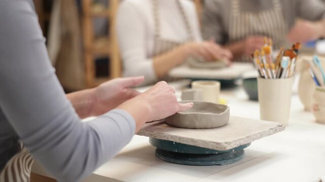 Close up unrecognizable female master forms bowl of clay during master class. Team of creative enthusiasts is engaged in clay modeling lesson in background