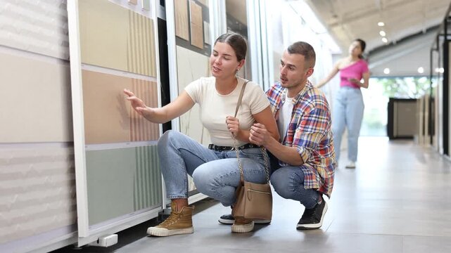 Interested young woman and man comparing ceramic tile samples displayed on racks in building hypermarket, while choosing materials for renovation to give home interior new, stylish look 