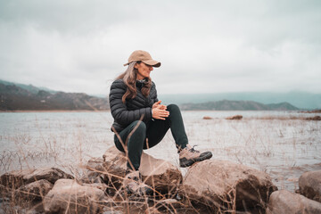 Woman in puffer jacket and hiking boots sits on rocks by a lake on a foggy day © Jezabel
