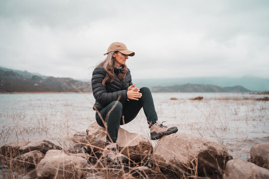 Woman in puffer jacket and hiking boots sits on rocks by a lake on a foggy day