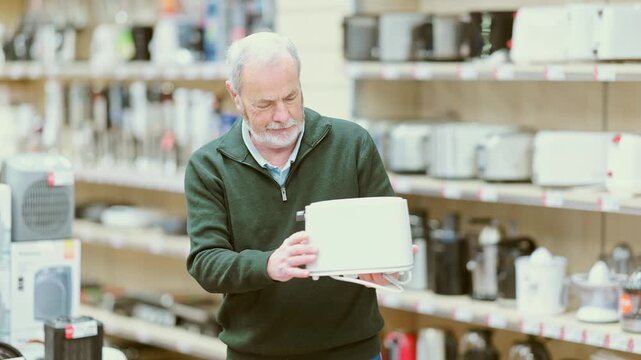 Mature man buyer examine toaster in detail before buying, checks completeness of household appliances and presence of defects. Customer chooses household appliances in store exhibition hall