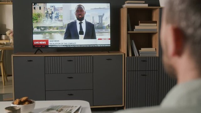 Over-shoulder shot of anonymous male televiewer turning on cable TV channel with remote control to watch war news, of soldiers running into combat, explosions, smoke, firing weapons