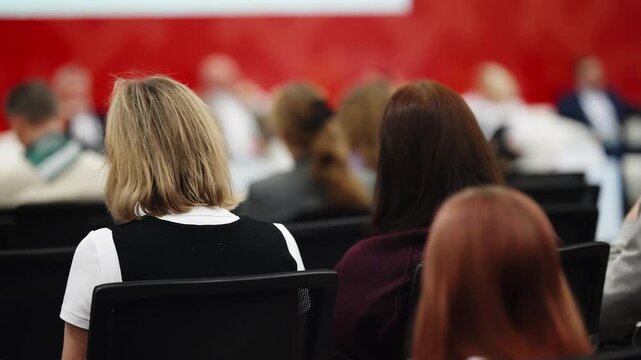 Rear view of attendees seated in rows, listening to a panel discussion at a professional conference, with blurred speakers on stage against a red backdrop in a formal setting