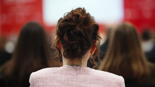 Back view of a woman with a curly updo in a pink blazer attentively listening among a blurred audience during a professional conference presentation in a red-accented venue