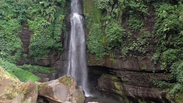 A cinematic drone shot capturing a serene waterfall flowing into a rocky gorge, nestled within the dense, lush green rainforest of East Java, Indonesia